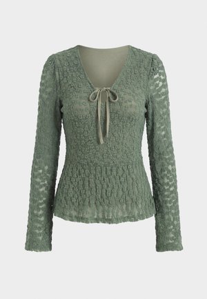 Blouse verte texturée à manches longues avec col en V et lien à l’avant, présentant une taille légèrement froncée et un motif de tissu semi-transparent.