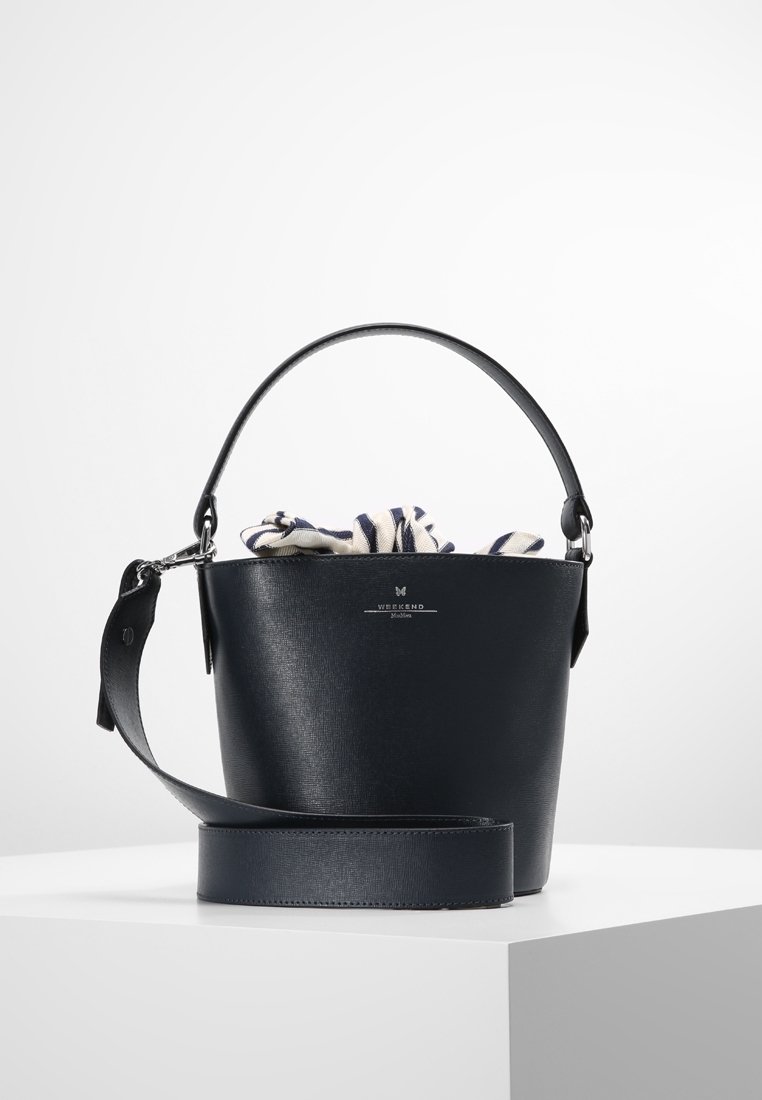 Sac seau en cuir noir avec poignée supérieure et bandoulière large amovible, partiellement rempli d'un tissu rayé à l'intérieur.