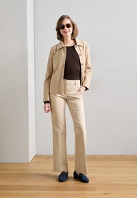 Blazer beige con botones, top ajustado marrón, pantalones beige de corte entallado y zapatos de estilo mocasín azul marino, complementados con gafas de sol oscuras. Fondo de suelo de madera.
