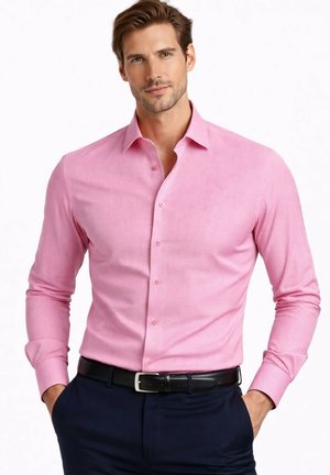 Homme portant une chemise rose cintrée à boutons, rentrée dans un pantalon bleu marine foncé avec une ceinture noire, mains dans les poches, fond neutre.