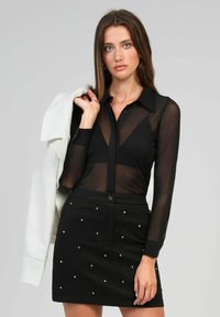 Blouse noire transparente à boutons avec des manches longues, associée à une mini-jupe noire ornée d'accents blancs en forme de perles.
