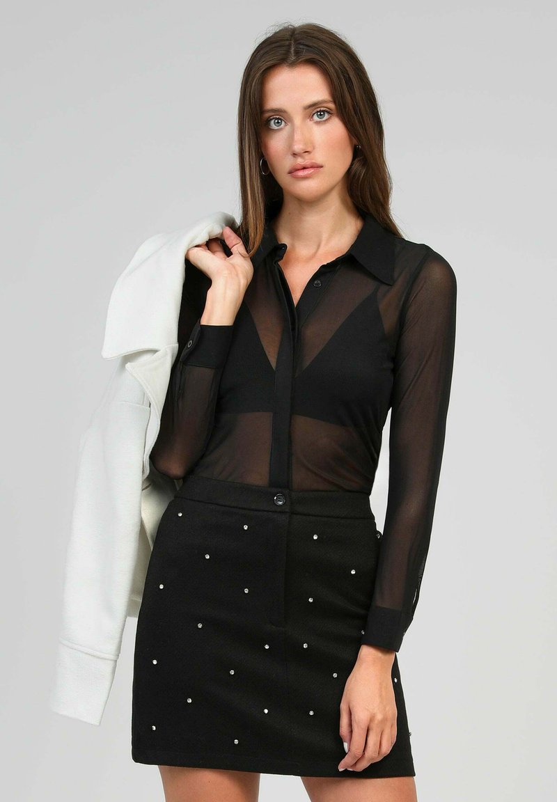 Blouse noire transparente à boutons avec des manches longues, associée à une mini-jupe noire ornée d'accents blancs en forme de perles.