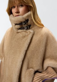 Poncho in misto lana beige con colletto alto, dotato di due cinghie in pelle marrone e fibbie in argento. Tessuto morbido e testurizzato con dettagli a costine.