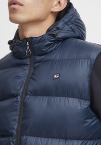 Mörkblå puffer väst med huva, som har en framtill dragkedja, justerbara snoddar och en liten logotyp på bröstet. Slät, glänsande yta.