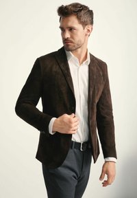 Ciemnobrązowa skórzana marynarka o dopasowanym kroju, z pojedynczym zapięciem na guzik i klapami. Nosi się ją na białej koszuli, w połączeniu z szarymi spodniami.