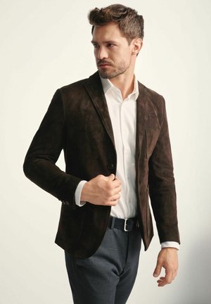 Ciemnobrązowa skórzana marynarka o dopasowanym kroju, z pojedynczym zapięciem na guzik i klapami. Nosi się ją na białej koszuli, w połączeniu z szarymi spodniami.