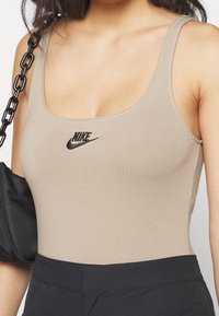 Beige, åtsittande linne gjort av mjukt tyg, med en svart Nike-logotyp i mitten. Kombineras med högmidjade svarta byxor.