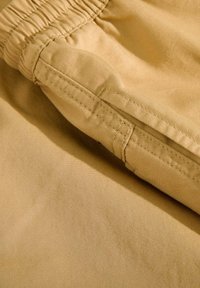 Tela marrón con textura suave, cinturilla elástica y costuras cosidas. Presenta un bolsillo plano en la pierna. Vista en primer plano del material.