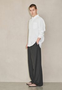 Jeune homme debout à l'intérieur, portant une chemise blanche à boutons, un pantalon noir ample et des sandales foncées, sur un fond neutre.