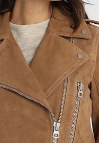 Hnědá nubuková motocyklová bunda se stříbrnými detaily, asymetrickým předním zipem a druky. Měkká textilie a detail nakloněného límce.