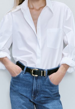 Woman wearing a white button-up shirt with rolled sleeves and blue jeans with a black belt, hands in pockets, against a plain background.