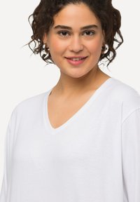 Weißes V-Ausschnitt-T-Shirt aus weichem Stoff, mit lockerem Schnitt und kurzen Ärmeln. Das Modell hat lockige Haare und trägt runde Ohrringe.