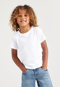 Vit bomulls-t-shirt med kort ärm och rund halsringning, bärs tillsammans med blå denimjeans. Barnet ler, händerna i fickorna.