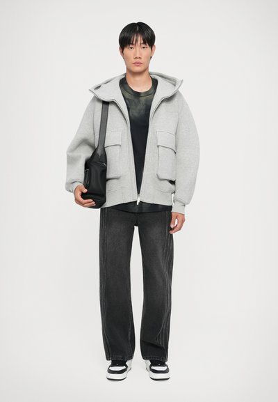 Un sweat à capuche gris zippé avec de grandes poches avant, superposé sur un t-shirt graphique noir. Associé à un jean large foncé et des baskets noir et blanc.