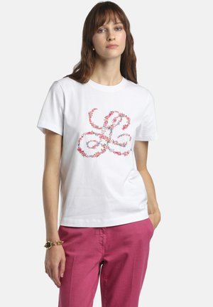 Nő fehér pólóban, rajta színes virágmintás spirál dizájn és rózsaszín nadrágban, egyik kezét a zsebében tartva, fehér háttér előtt áll.