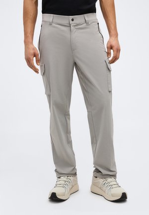 Mand iført lysegrå cargobukser med klaplommer og beige sportssko, stående mod en ensfarvet baggrund.