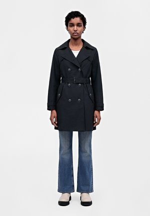 Le modèle se tient face à l’objectif, vêtu d’un manteau noir croisé et ceinturé, d’un jean bleu et de chaussures noires et blanches, sur un fond clair uni.