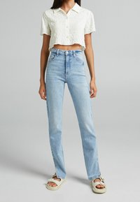 Cropped, white, textured shirt with short sleeves, paired with high-waisted, light blue, straight-leg jeans. Casual footwear completes the outfit.