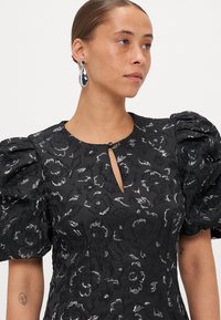 Čierne šaty s textúrovaným kvetinovým vzorom, pufové rukávy a detail výstrihu s kľúčovou dierkou. Strieborné doplnky dopĺňajú vzhľad.