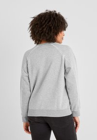 Woman with curly hair wearing a light gray sweatshirt with decorative stitching on sleeves and back, paired with black jeans, facing away.