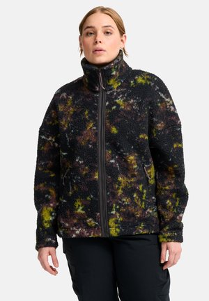Junge Person trägt eine Fleecejacke mit hohem Kragen und Reißverschluss mit einem schwarz-gelb-braunen Batikmuster und schwarze Hose vor weißem Hintergrund.