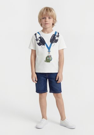 Blonder Junge, der ein weißes T-Shirt mit aufgedruckten Kopfhörern und grünem Dinosaurier-Schlüsselanhänger trägt, Jeans-Shorts und weiße Turnschuhe, stehend.