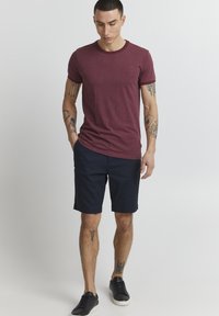 T-shirt bordeaux à col rond, associé à un short bleu marine. Le modèle porte des baskets noires et a des tatouages visibles sur les bras et les jambes.
