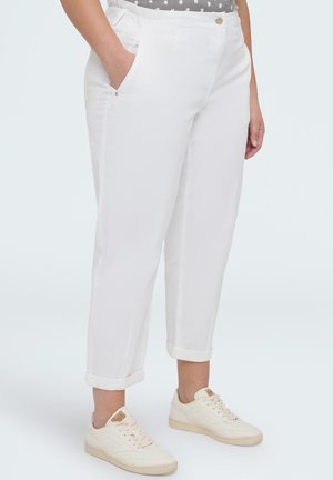 Person iført hvide bukser med opkrogede ben, en mønstret indstoppet top og lyse beige sneakers, stående med den ene hånd i lommen.