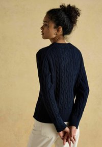 Žena s kudrnatými vlasy spletenými do drdolu, oblečená v námořnické modré svetr s pleteným vzorem a smetanových kalhotách, stojící s rukama zkříženýma za zády.