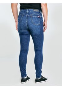 Inimene, kes kannab siniseid kitsaid teksaseid ja musti tossusid, seisab seljaga kaamera poole valge tavalise tausta ees.