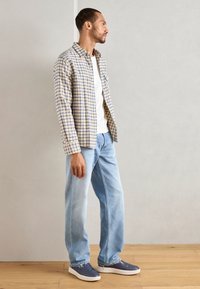 Chemise à carreaux jaune et bleue, t-shirt blanc en dessous, jean bleu clair et baskets grises avec semelles blanches. Debout sur un sol en bois.