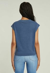 Žena s dlouhými zapletenými vlasy, oblečená v bezrukávovém modrém topu a světlých modrých džínách, stojí zády k fotoaparátu před jednobarevným pozadím.