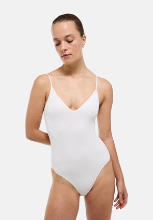 Jeune femme aux cheveux attachés, portant un maillot de bain blanc une pièce sans manches sur un fond clair uni.