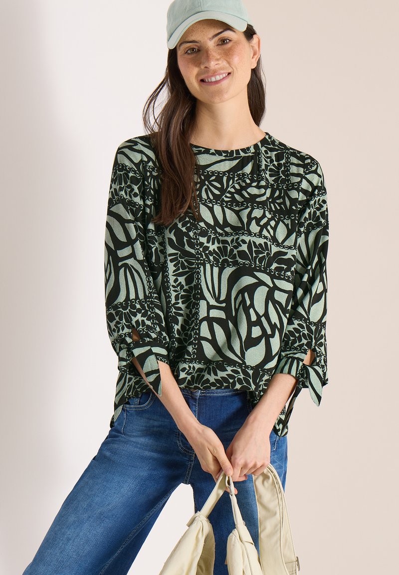 Femme portant un haut à manches longues à motifs verts, un jean bleu et une casquette vert clair, tenant un sac beige à deux mains devant elle.