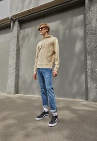Beige hettegenser med frontlomme og snøring, blå jeans og svarte joggesko med hvite såler og snørebånd. Herre uformell antrekk.