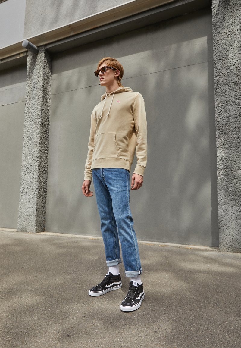 Beige hettegenser med frontlomme og snøring, blå jeans og svarte joggesko med hvite såler og snørebånd. Herre uformell antrekk.