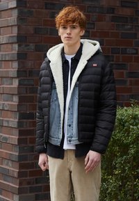 Chaqueta de plumas negra con capucha forrada de piel sintética, superpuesta a una chaqueta de mezclilla envejecida, combinada con pantalones de pana beige. Paleta de colores neutros.