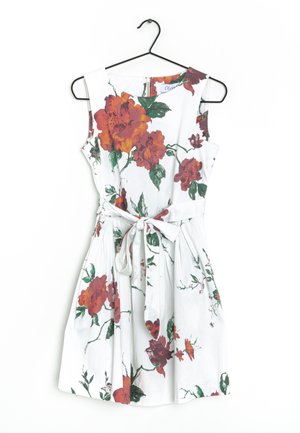 Hvid ærmeløs kjole med store røde og grønne blomsterprint, med bundet talje og synligt lynlåsdetalje på ryggen. Hænger på en sort krog.