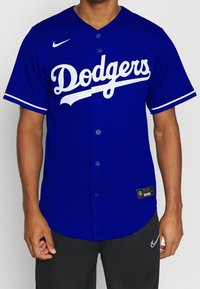 Modrá baseballová dres s bílým zakřiveným nápisem "Dodgers", zapínání na knoflíky vpředu a krátké rukávy. Logo Nike na pravé straně hrudi.