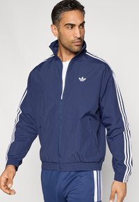 Tmavomodrá športová bunda z ľahkého materiálu, so zipsom vpredu, vysokým golierom a bielymi tromi pruhmi na rukávoch. Na hrudi má logo.