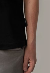 Schwarzes T-Shirt mit kurzen Ärmeln aus strukturiertem Stoff, mit sichtbarem Markenschild an der Seite, kombiniert mit hellgrauen Hosen.