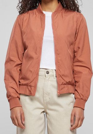 Personne portant une veste bomber couleur rouille par-dessus une chemise blanche et un pantalon beige taille haute sur un fond uni.