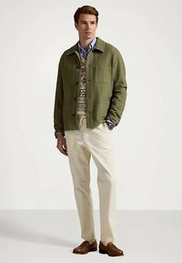 Man som bär en grön jacka över en mönstrad tröja och blå skjorta, vita byxor och bruna loafers, står med en hand i fickan.