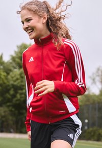 Red zip-up jacket with white accents and three white stripes on each sleeve, paired with black shorts. Fabric appears lightweight and sporty.