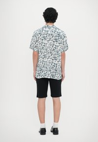 Κοντομάνικο πουκάμισο σε λευκό χρώμα με μπλε φυτικό σχέδιο, συνδυασμένο με μαύρες βερμούδες και κάλτσες. Σκούρα παπούτσια συμπληρώνουν το σύνολο.