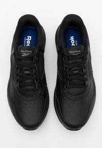 Chaussures de sport noires avec une tige en cuir, fermeture à lacets et accents gris texturés. Intérieur doté d'une semelle intérieure bleue portant l'inscription "Reebok".