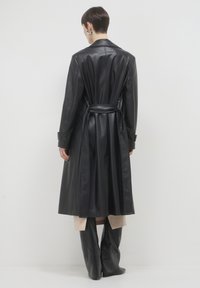 Trench coat in pelle nera con cintura, maniche lunghe e polsini con bottoni. Tessuto liscio, vestibilità su misura, design lungo fino al ginocchio.