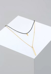 Gouden ketting met een lange, slanke hanger aan een fijne ketting, gelaagd over een kortere zwarte draad met een driehoekige gouden charme.