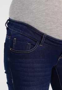 Jeans de mezclilla azul oscuro con bolsillos frontales, costuras en contraste y una cinturilla alta, combinados con una blusa gris claro. La textura parece suave.