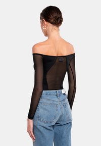 Schwarzer Off-Shoulder-Bodysuit mit langen durchsichtigen Mesh-Ärmeln, figurbetonter Silhouette und einem Etikett am Rücken, kombiniert mit hochgeschnittenen blauen Jeans.